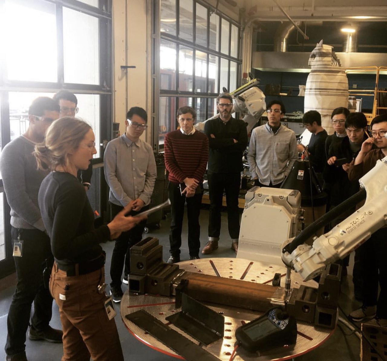 Stefanie Pender explaining a robotic welding cell to a group in the shop, teach pendant and fixture table in the foreground