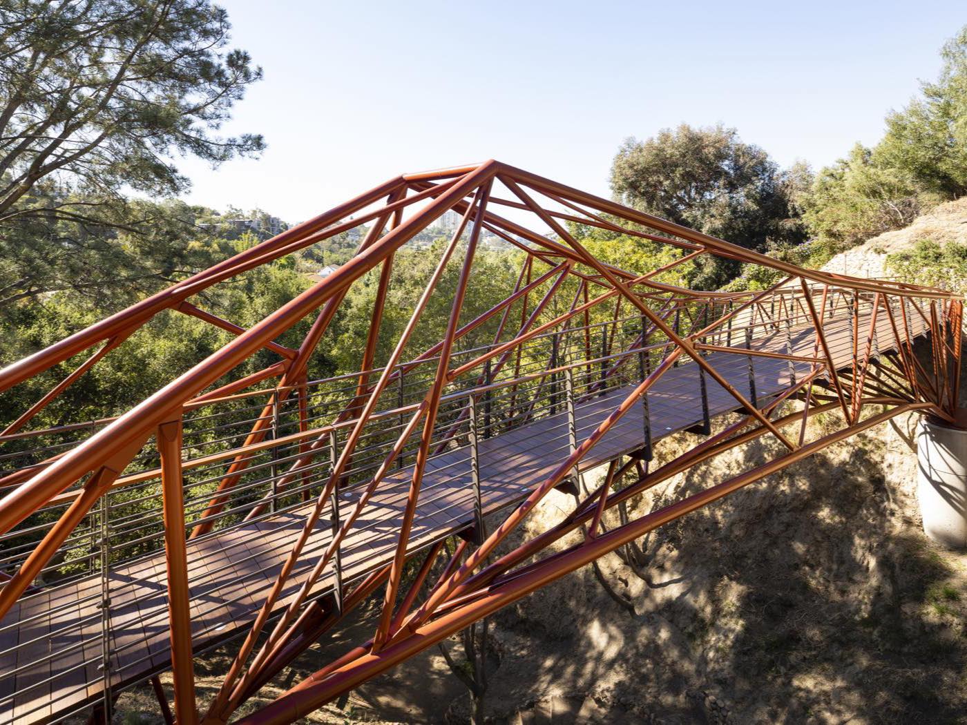 The completed Arroyo Bridge, an orange steel lattice pedestrian bridge spanning a wooded canyon in Los Angeles