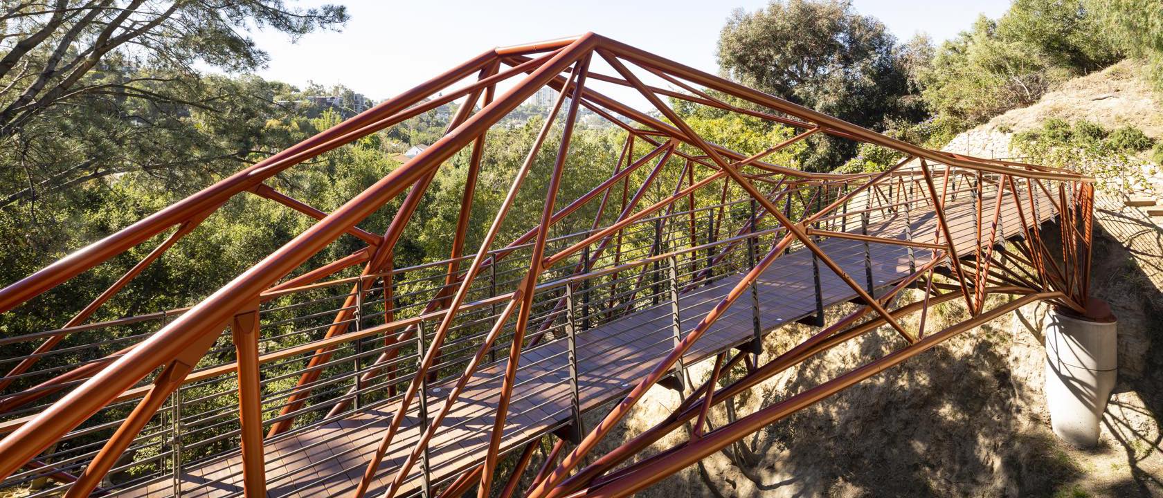 The completed Arroyo Bridge, an orange steel lattice pedestrian bridge spanning a wooded canyon in Los Angeles