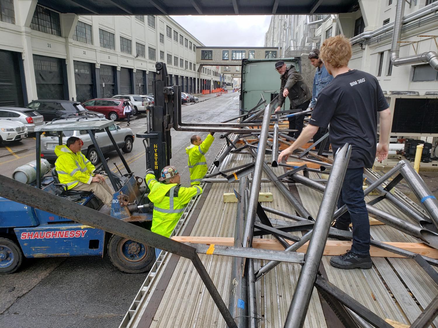 Workers and a forklift loading welded steel lattice sections of the bridge onto a flatbed truck outside the shop