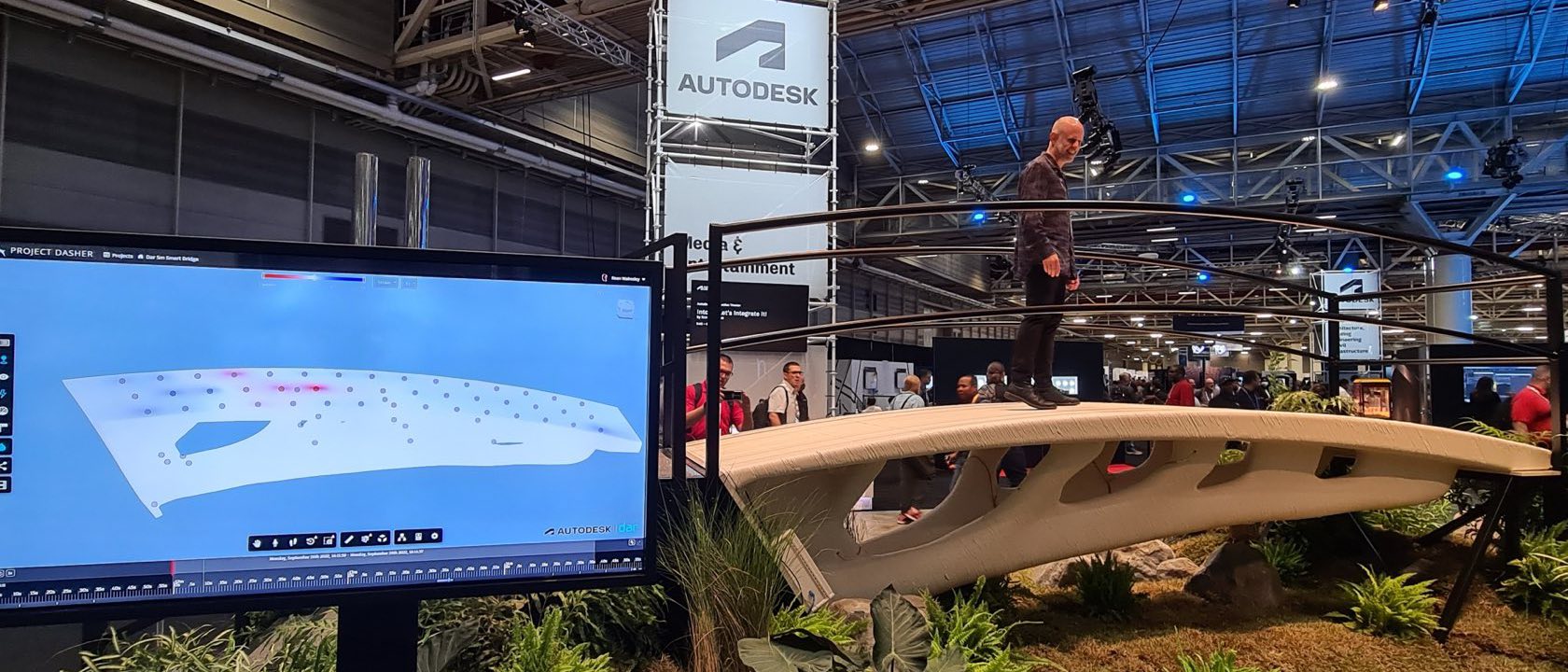 A person walking across the printed bridge on an exhibition floor, with a live digital-twin readout of the structure on a screen beside it