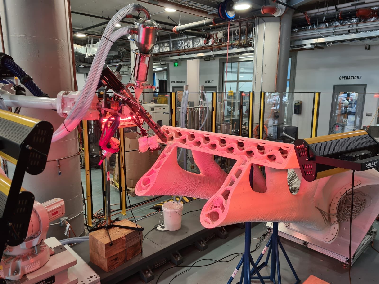 An ABB robot arm with a pellet extruder printing the bridge structure, lit in red by the heated end effector