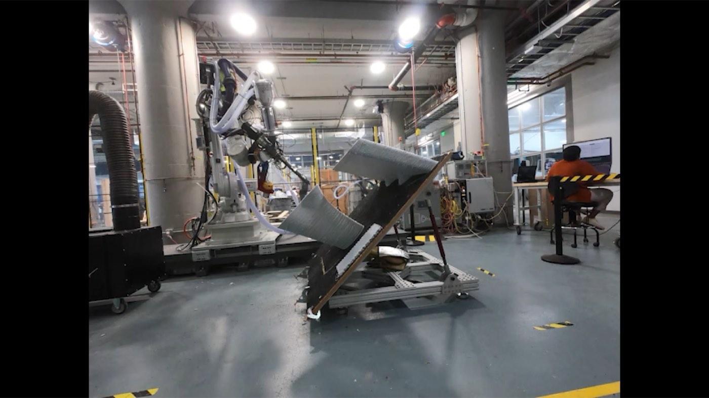 An ABB robot printing onto a steeply angled print bed in the shop, with an operator monitoring from a workstation