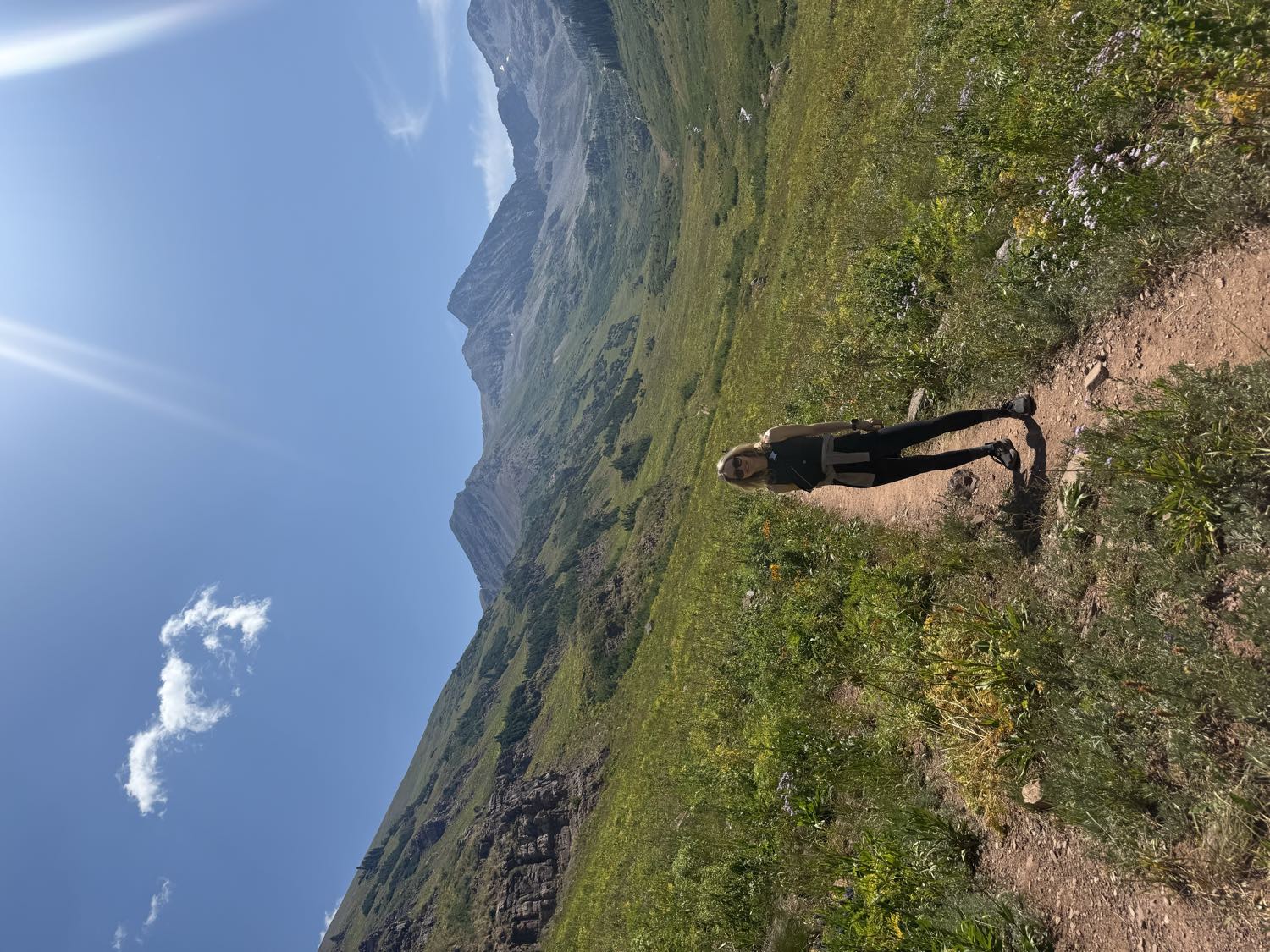 Stefanie Pender standing on a trail in an alpine valley with wildflowers and mountain peaks behind