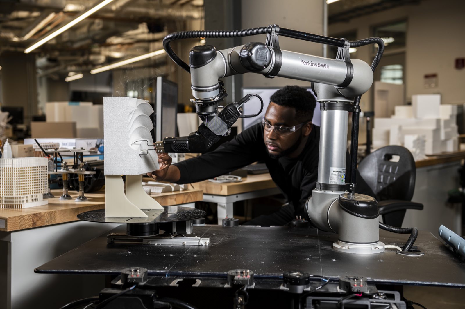 A researcher guiding a Universal Robots collaborative arm shaping a foam architectural model on a rotary table