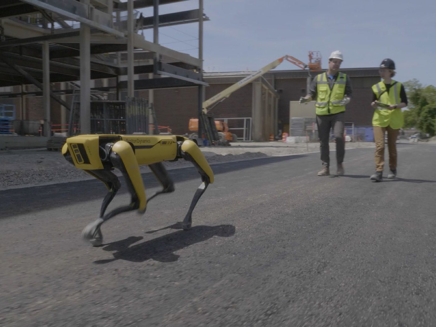 A yellow Boston Dynamics Spot quadruped robot walking across fresh asphalt on an active construction site, with two workers in hi-vis vests following behind