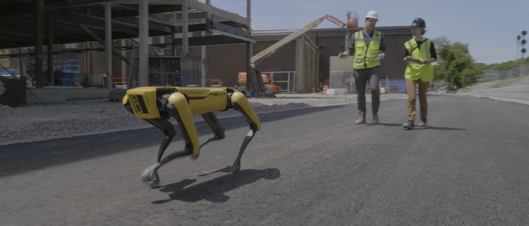 A yellow Boston Dynamics Spot quadruped robot walking across fresh asphalt on an active construction site, with two workers in hi-vis vests following behind