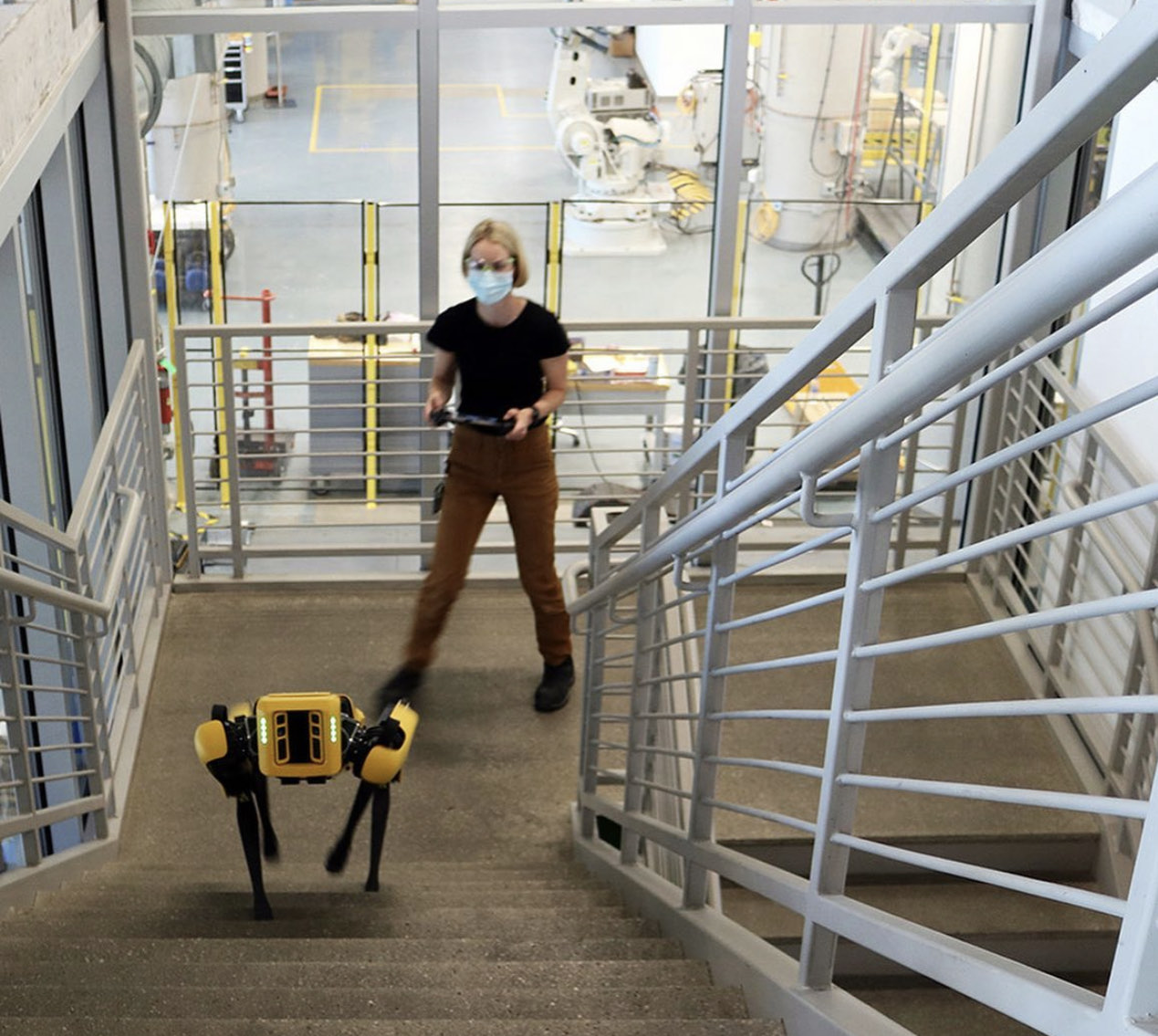 Stefanie Pender piloting the Spot robot up a staircase inside the robotics lab, controller in hand, with industrial robot cells on the floor below