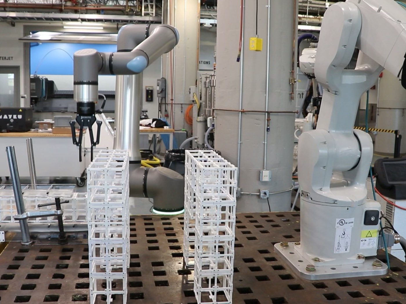 A Universal Robots arm and an ABB arm assembling white lattice voxel modules into towers on a steel fixture table