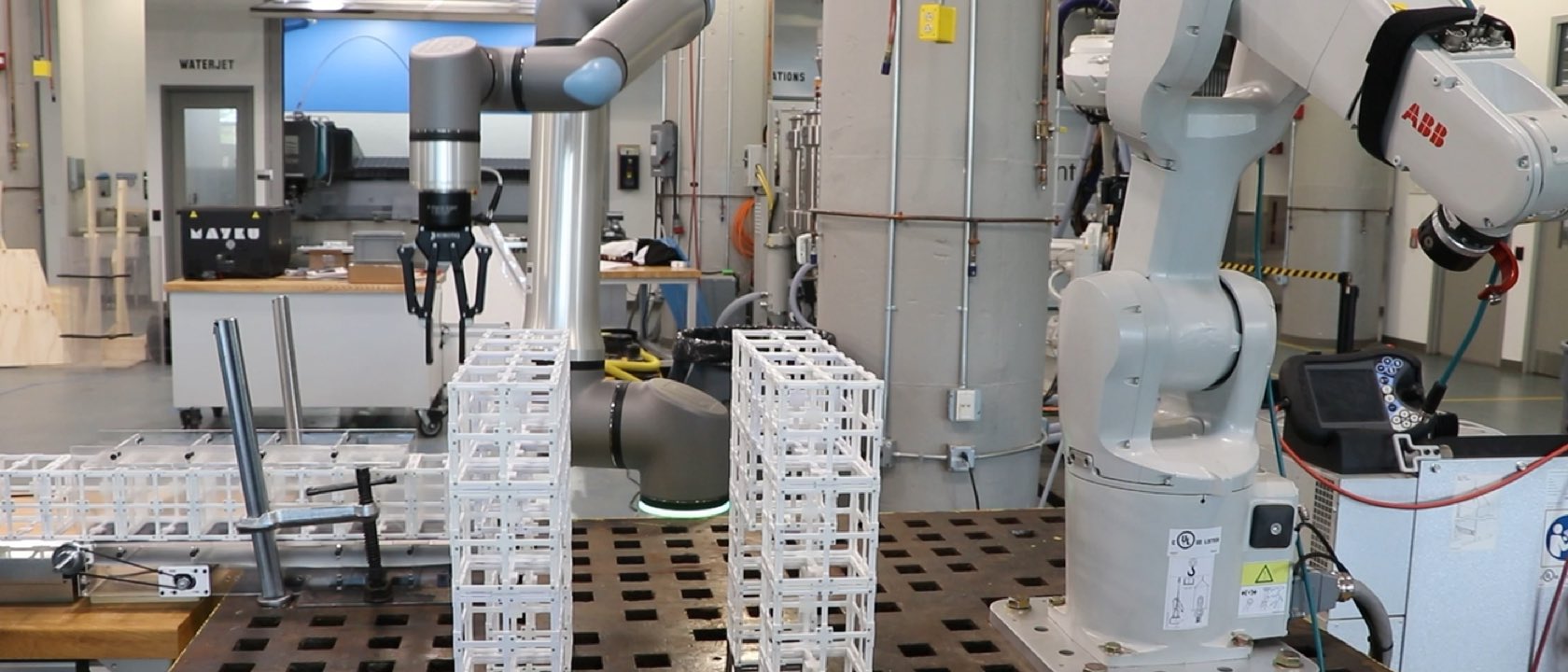 A Universal Robots arm and an ABB arm assembling white lattice voxel modules into towers on a steel fixture table